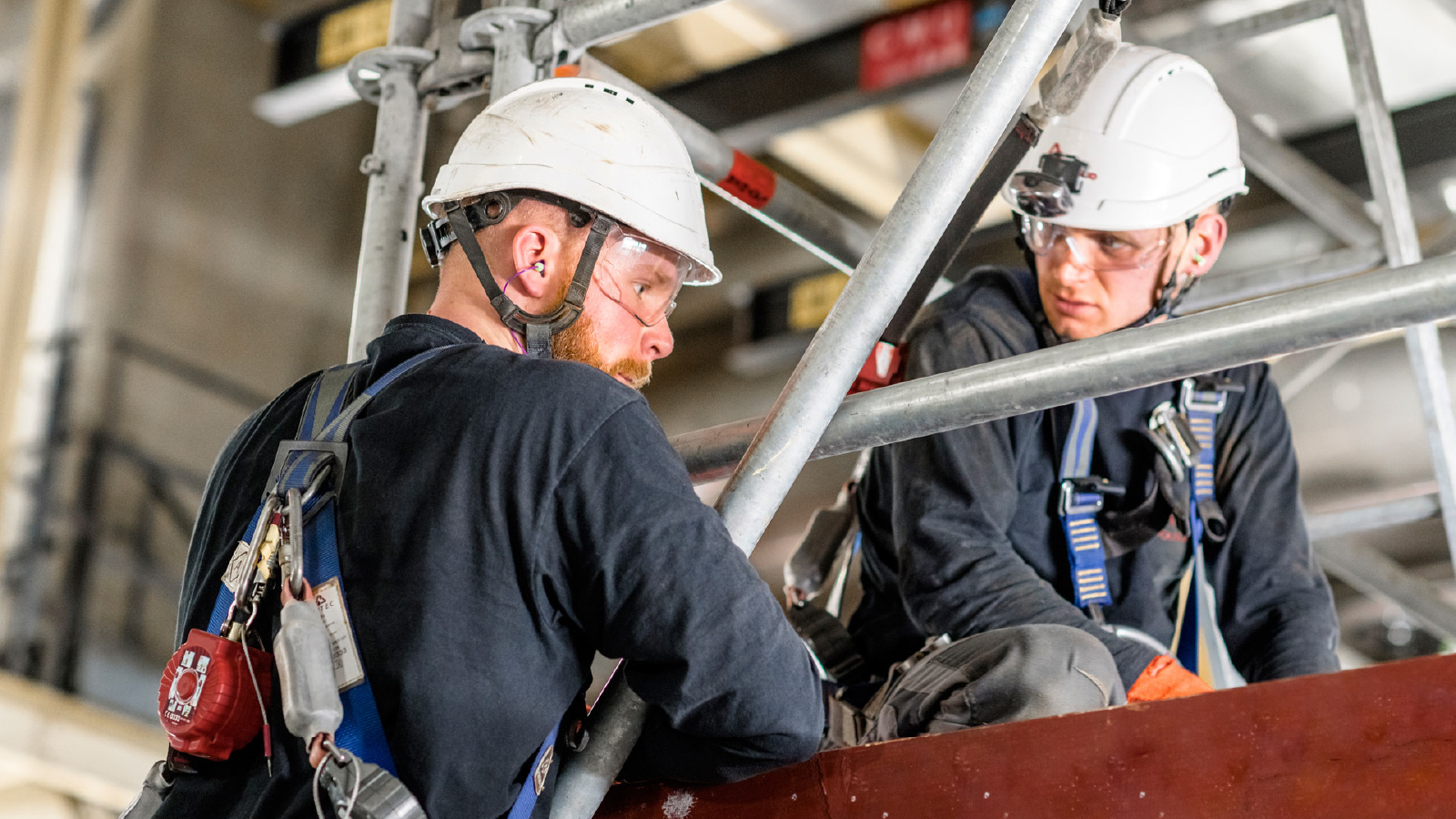 Two workers in black uniforms and white safety helmets with harnesses and protective gear conduct elevated maintenance on a steel scaffolding structure.