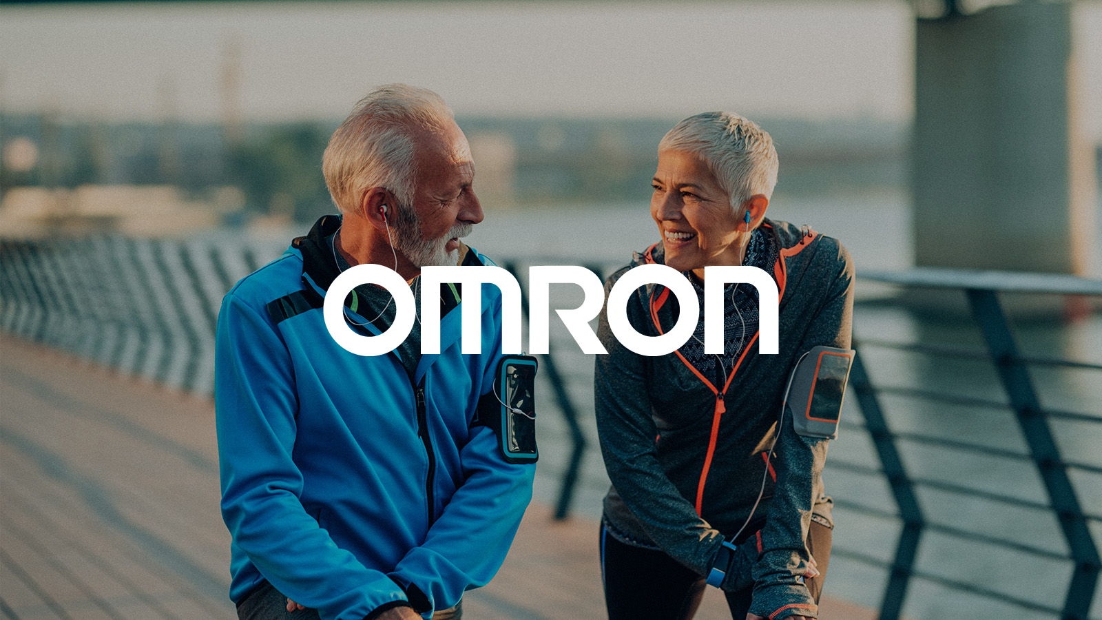 Omron white logo in a image of an older man and woman in athletic wear smiling and talking while resting outdoors with earphones and smart devices, suggesting active and healthy living.