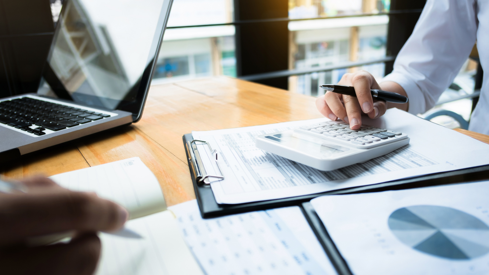 Hands of two cowrokers writing and using a calculator on a pile of reports and charts on a conference table